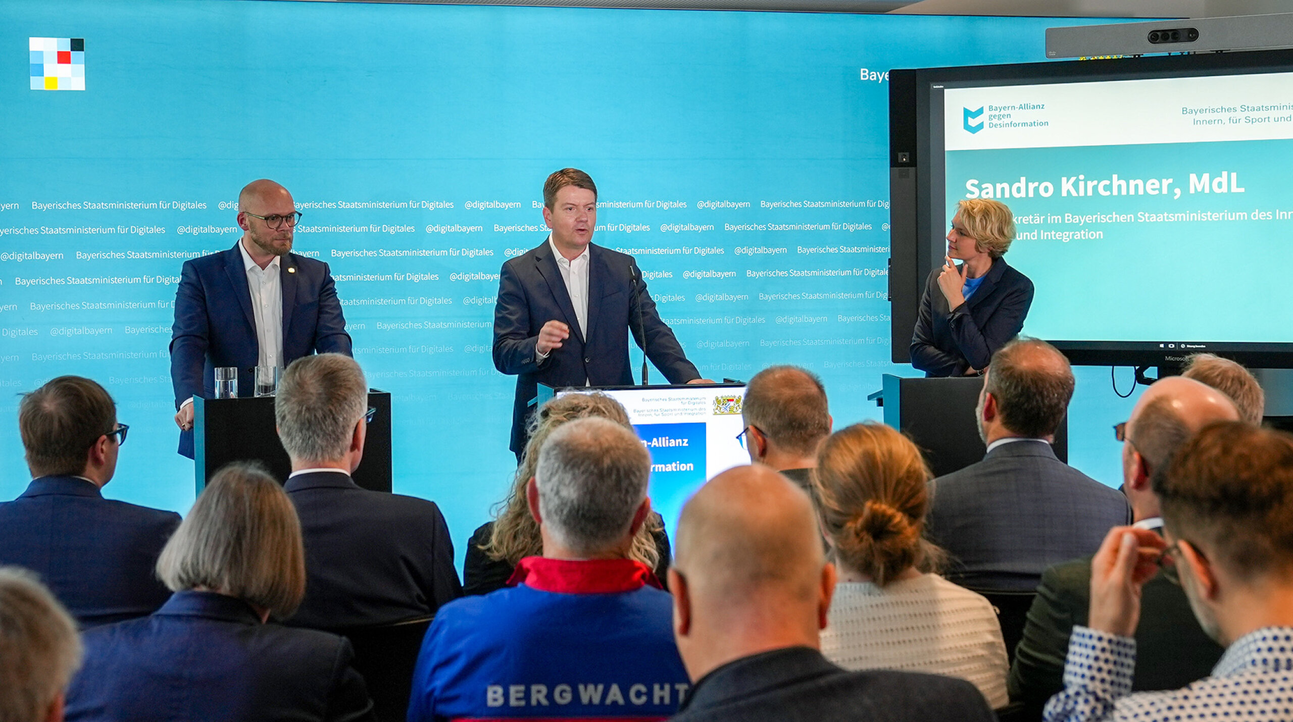 Staatssekretär Sandro Kirchner (rechts), Staatsminister Dr. Mehring (links) stehen vor einer Pressewand mit Blick zum Publikum - rechts steht eine Moderatorin