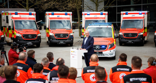 Herrmann am Rednerpult vor Rettungswägen, im Vordergrund Rettungskräfte in Uniform von hinten