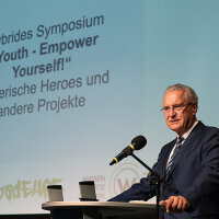 1. Juli 2021: Neues Integrationsprojekt „YeY! Youth – Empower Yourself!“: Innen- und Integrationsminister Joachim Herrmann gibt grünes Licht für die neue Dachmarke, unter die der Freistaat Bayern seine Vorzeigeprojekte in Sachen Gewaltprävention, Respekt und Toleranz bündelt. „Unsere Erfolgsprojekte „HEROES“, „habadEHRE“ und „W3“ leisten hier großartige Arbeit und tragen damit entscheidend zur kulturellen Integration junger Menschen mit Migrationshintergrund bei“, lobte Herrmann. Innen- und Integrationsminister Joachim Herrmann hinter Rednerpult vor Präsentationsleinwand
