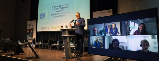 Innen- und Integrationsminister Joachim Herrmann gibt grünes Licht: Die neue Dachmarke „YeY! Youth – Empower Yourself!“ bündelt die Erfolgsprojekte in Sachen Gewaltprävention, Respekt und Toleranz. Innen- und Integrationsminister Joachim Herrmann hinter Rednerpult neben Bildschirm mit Videokonferenz