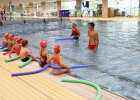 Kinder mit Schwimmnudel am Schwimmbeckenrand, Schwimmlehrer im Pool