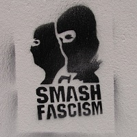 SMASH FASCISM Das Bild zeigt ein an eine Wand gesprühtes Graffiti. Dieses Graffiti in schwarzer Farbe zeigt zwei vermummte Gesichter und den Text „SMASH FASCISM“