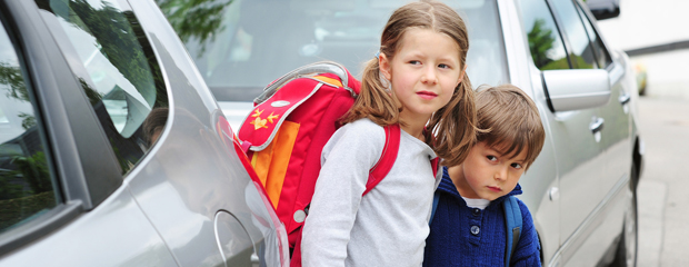 Sicher zur Schule - sicher nach Hause. Zwei Schulkinder stehen zwischen geparkten Autos.