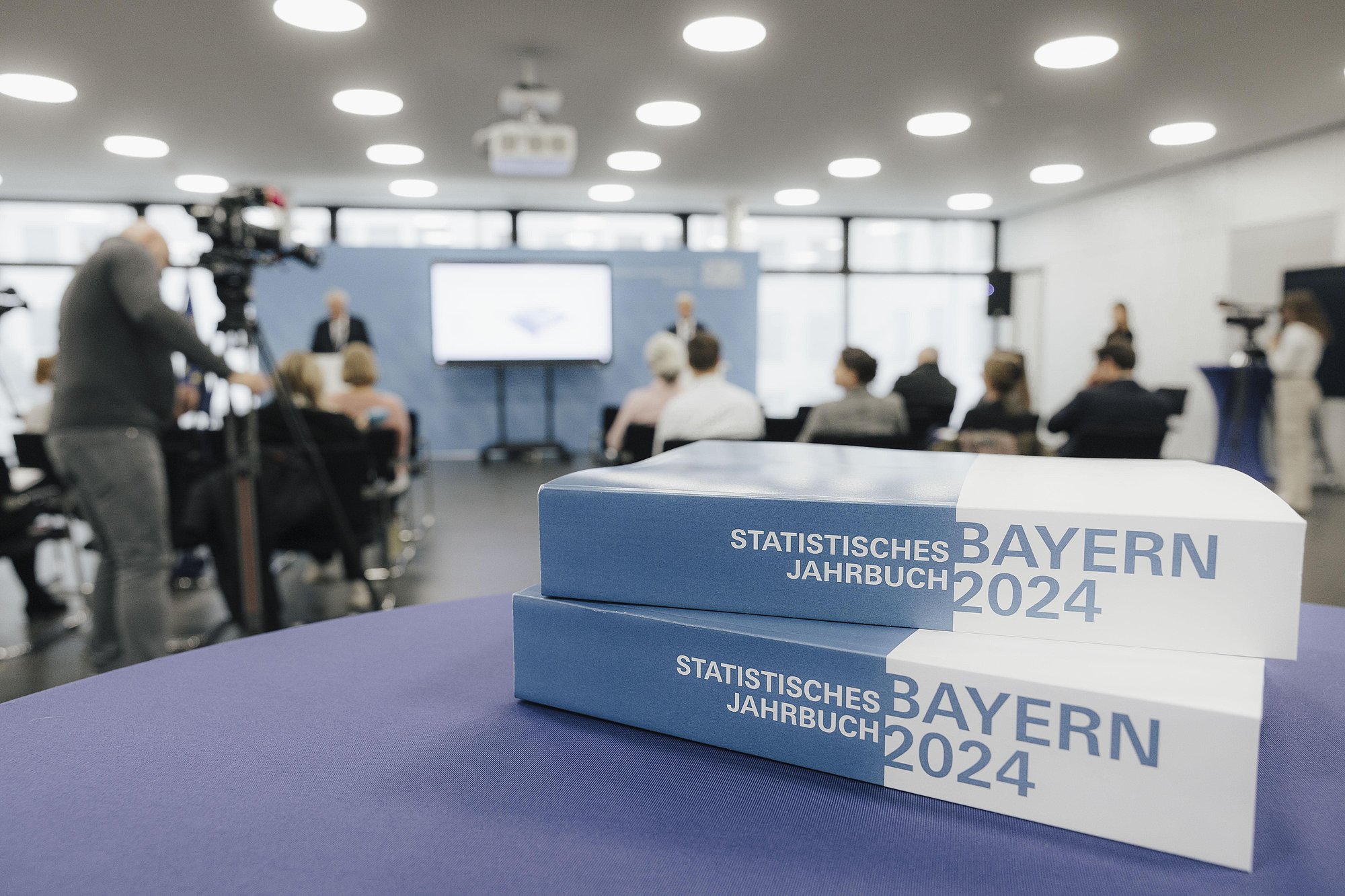 Zwei Ausgaben des Statistischen Jahrbuchs liegen auf einem Tisch, im Hintergrund findet eine Pressekonferenz statt 
