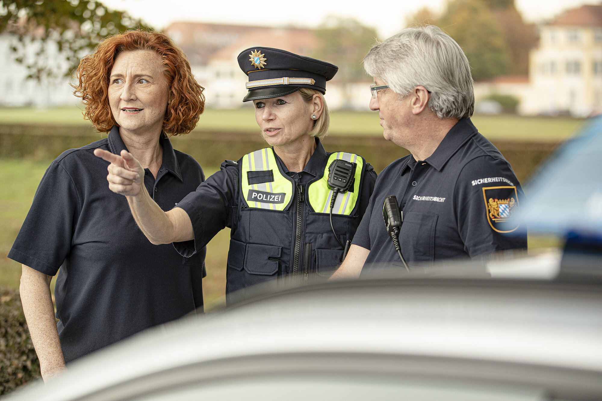 Polizistin mit zwei Ehrenamtlichen der Bayerischen Sicherheitswacht vor einem Auto. Sie schauen alle in eine Richtung, auf die die Polizistin hinweist