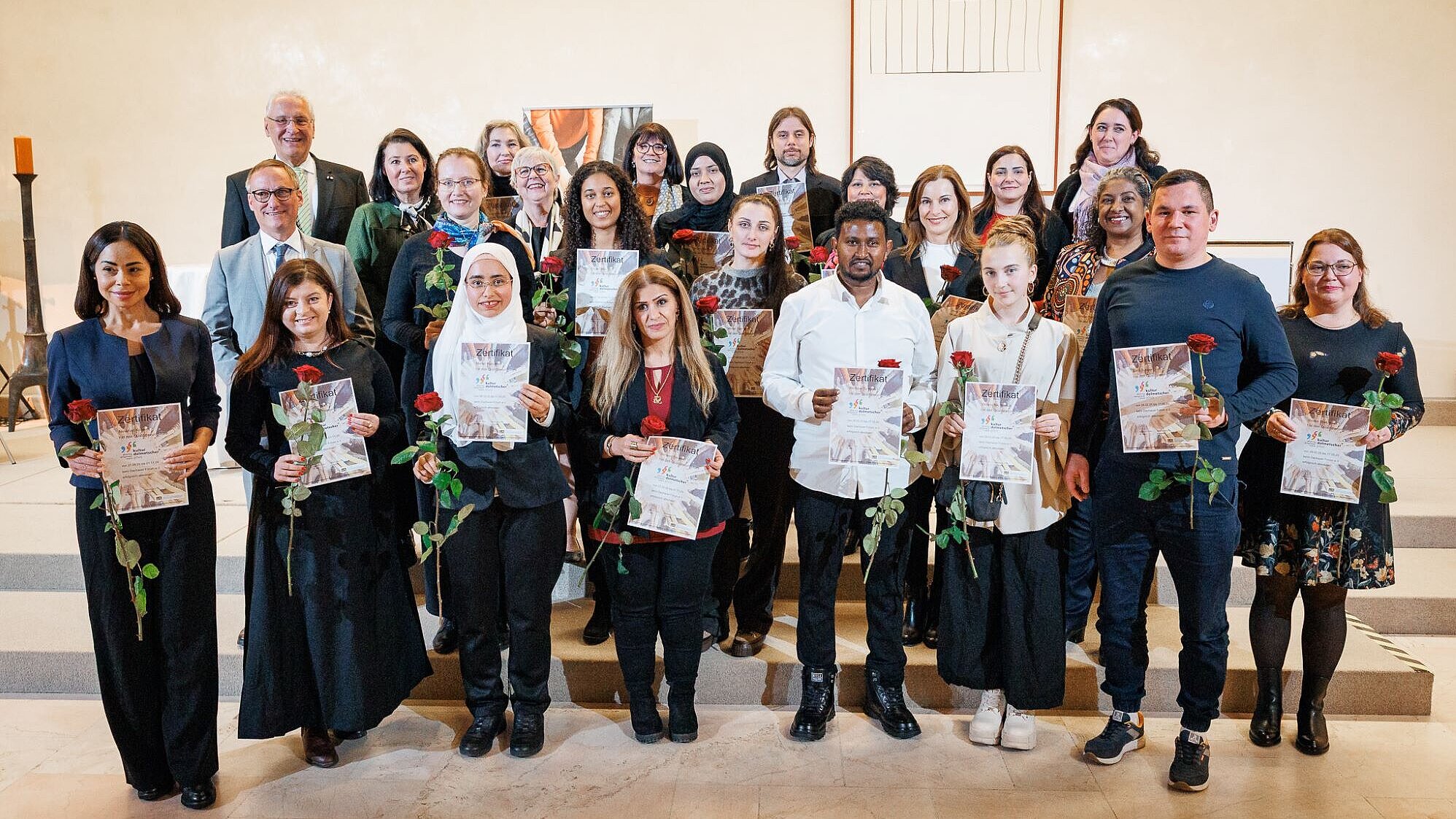 Ein Gruppenbild mit Innenminister Joachim Herrmann und allen Kulturdolmetscherinnen und Kulturdolmetschern. Alle haben ihre Urkunde sowie eine Rose in der Hand und blicken zum Fotografen.