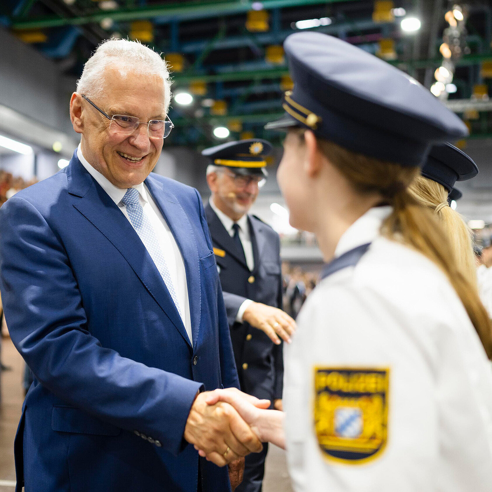 Begrüßungsszene in einer großen Halle: Ein Mann in blauem Anzug reicht einer jungen Beamtin in weißer Polizeiuniform die Hand; im Hintergrund stehen weitere uniformierte Personen in Reihen.