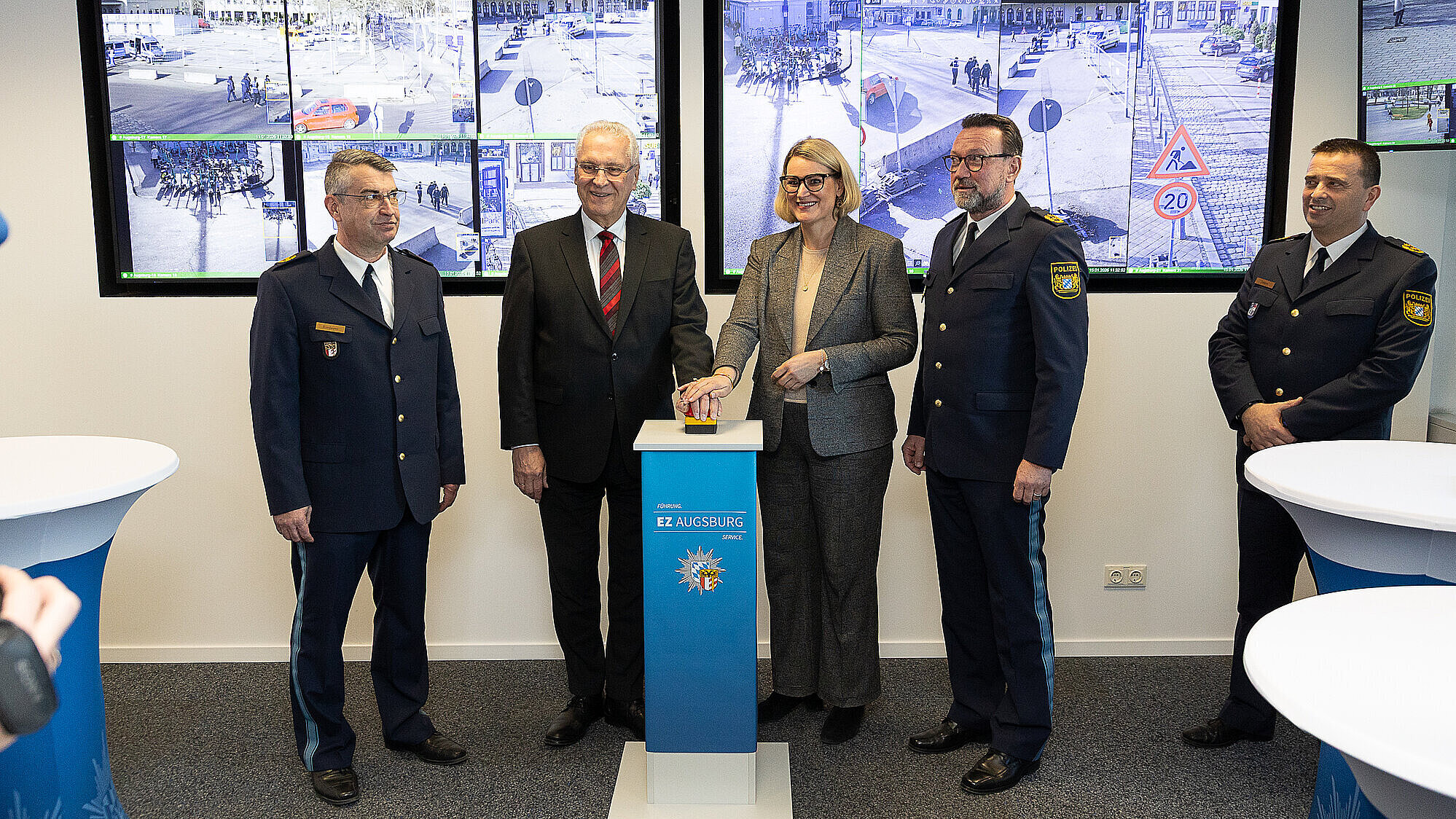 Innenminister Joachim Herrmann mit weiteren Personen vor zwei Bildschirmen, die öffentliche Straßen zeigen. Die Personen halten ihre Hand auf einen großen Knopf auf einem Podest. 