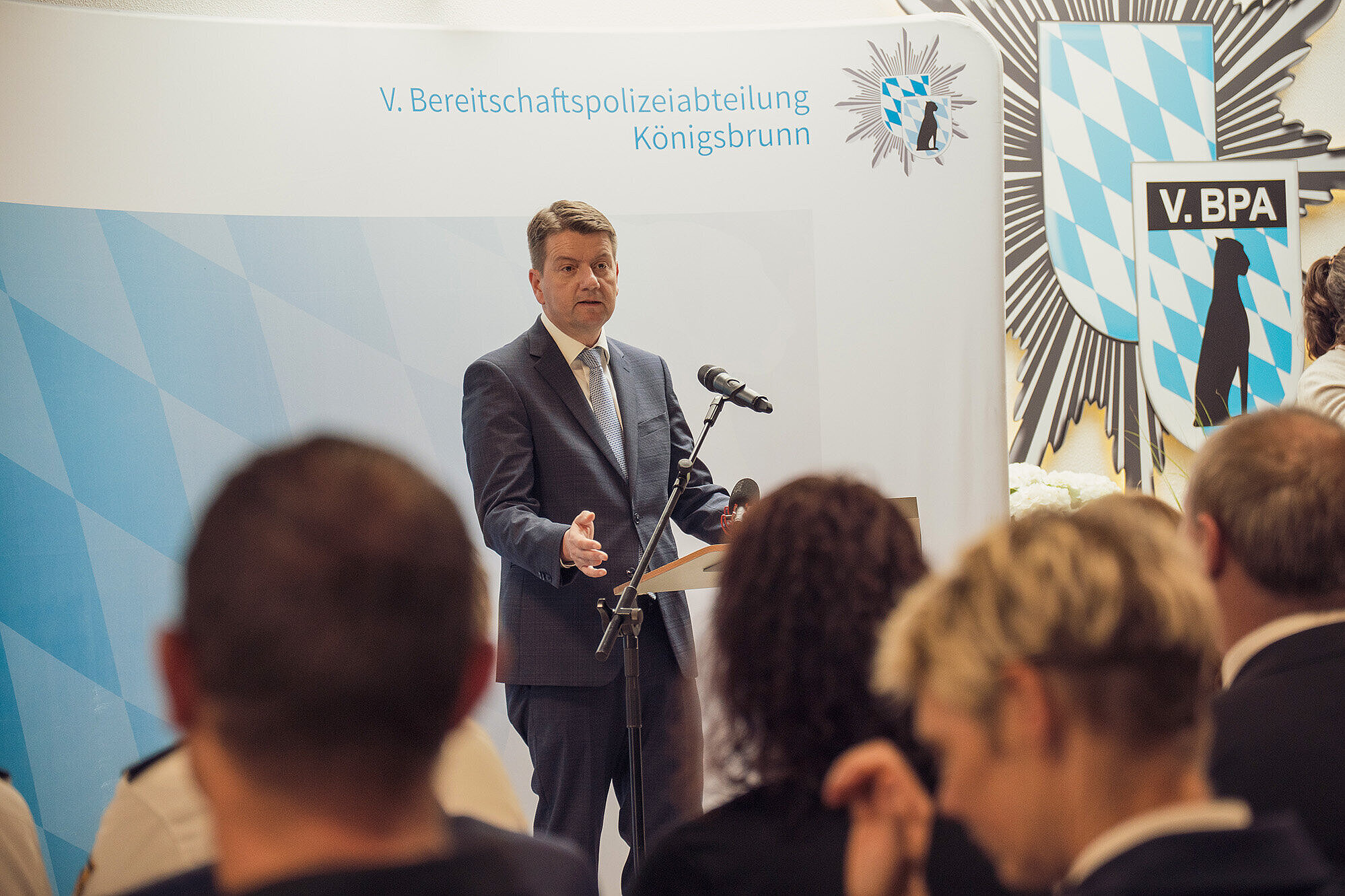 Innenstaatssekretär Sandro Kirchner in einem dunklen Anzug, der am Rednerpult steht und eine Rede hält. Im Hintergrund sind Wappen und Symbole der Bereitschaftspolizei Königsbrunn zu sehen, darunter das Logo der V. Bereitschaftspolizeiabteilung. Vor ihm sitzen mehrere Zuhörerinnen und Zuhörer, die aufmerksam zuhören.