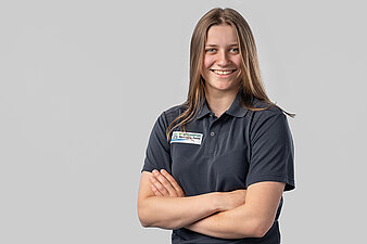 Leonie Bach-Staudinger vor hellem Hintergrund, trägt dunkles Poloshirt mit rechteckigem Aufnäher und hat die Arme verschränkt.
