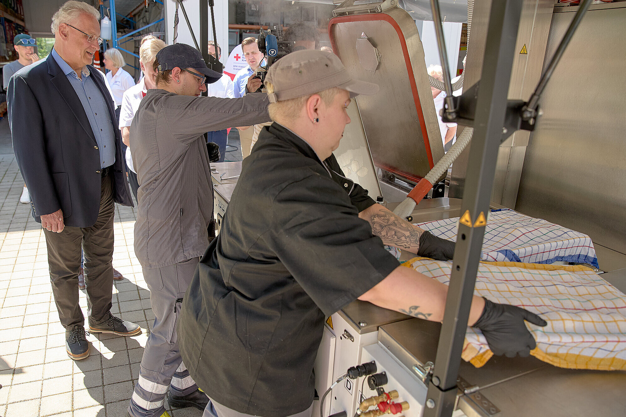 Innenminister Joachim Herrmann steht hinter einer Feldküche, an dieser zwei Köche arbeiten.