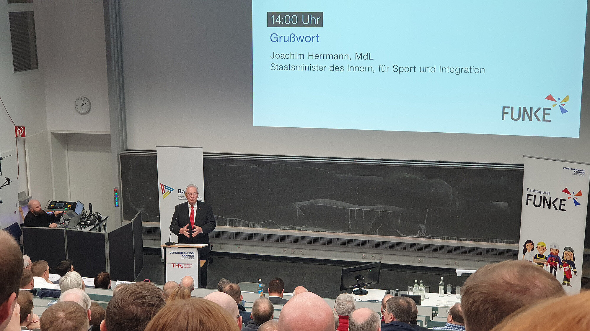 Eine Person in dunklem Anzug und roter Krawatte steht am Rednerpult in einem besetzten Hörsaal und hält eine Rede; auf der Projektionsleinwand oben ist das Programm mit dem Eintrag „14:00 Uhr – Grußwort“ zu sehen, rechts das FUNKE‑Logo.