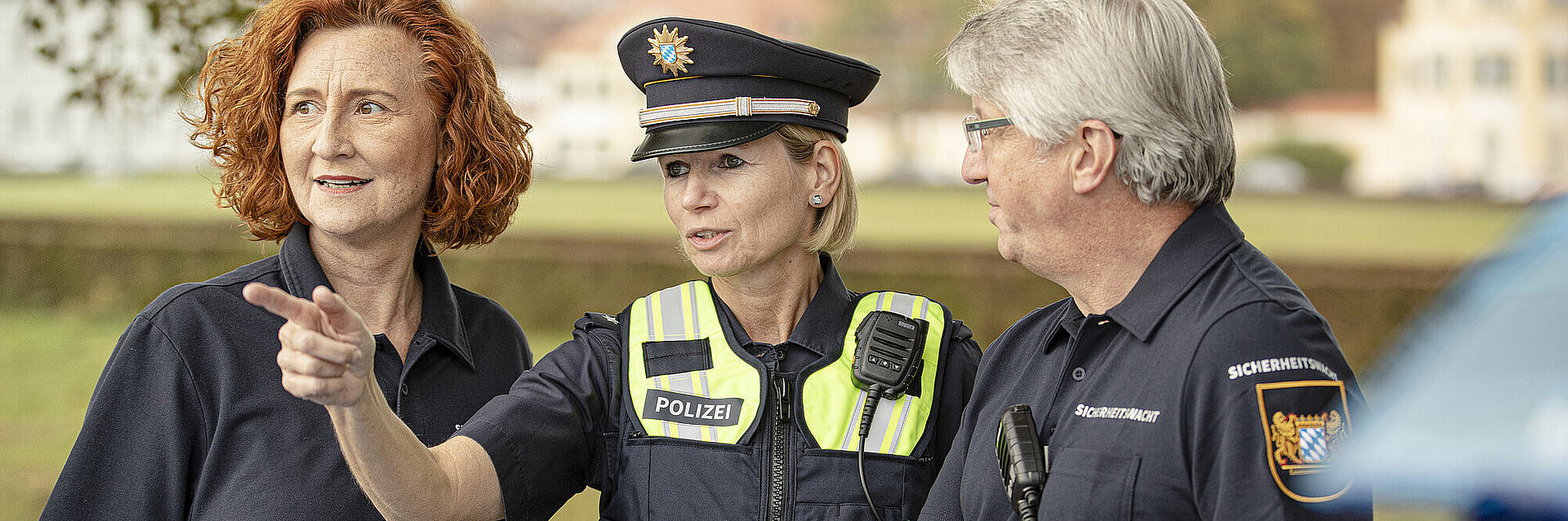 Polizistin mit zwei Ehrenamtlichen der Bayerischen Sicherheitswacht vor einem Auto. Sie schauen alle in eine Richtung, auf die die Polizistin hinweist