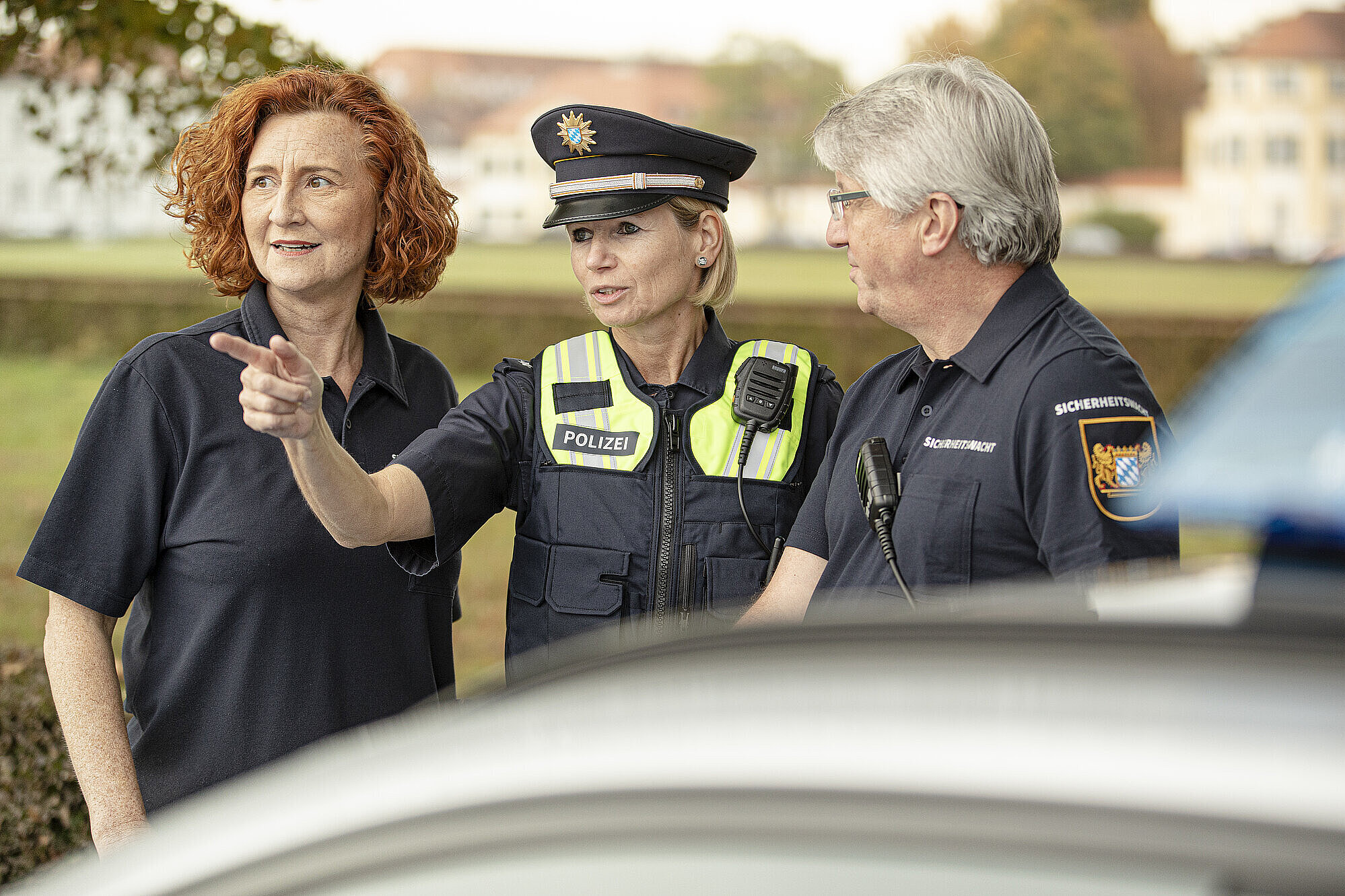 Polizistin mit zwei Ehrenamtlichen der Bayerischen Sicherheitswacht vor einem Auto. Sie schauen alle in eine Richtung, auf die die Polizistin hinweist