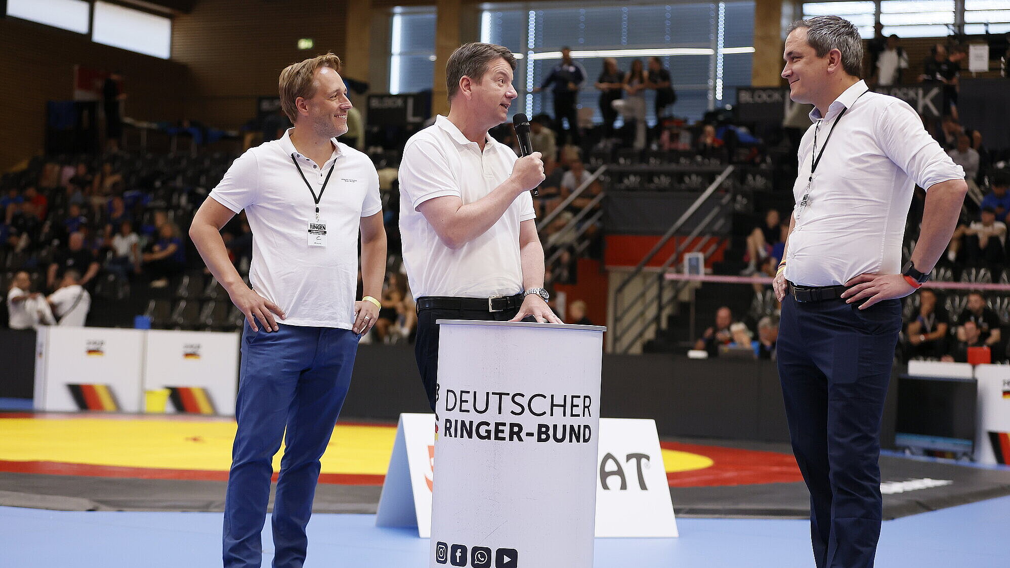 Kirchner mit Mikrofon bei Ansage neben zwei Männern in weißen Sportshirts in Sporthalle, im Hintergrund Tribüne