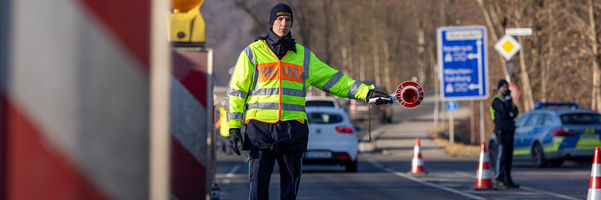 Polizist in Warnweste auf einer Straße mit Kelle, im Hintergrund weitere Polizisten und Autos