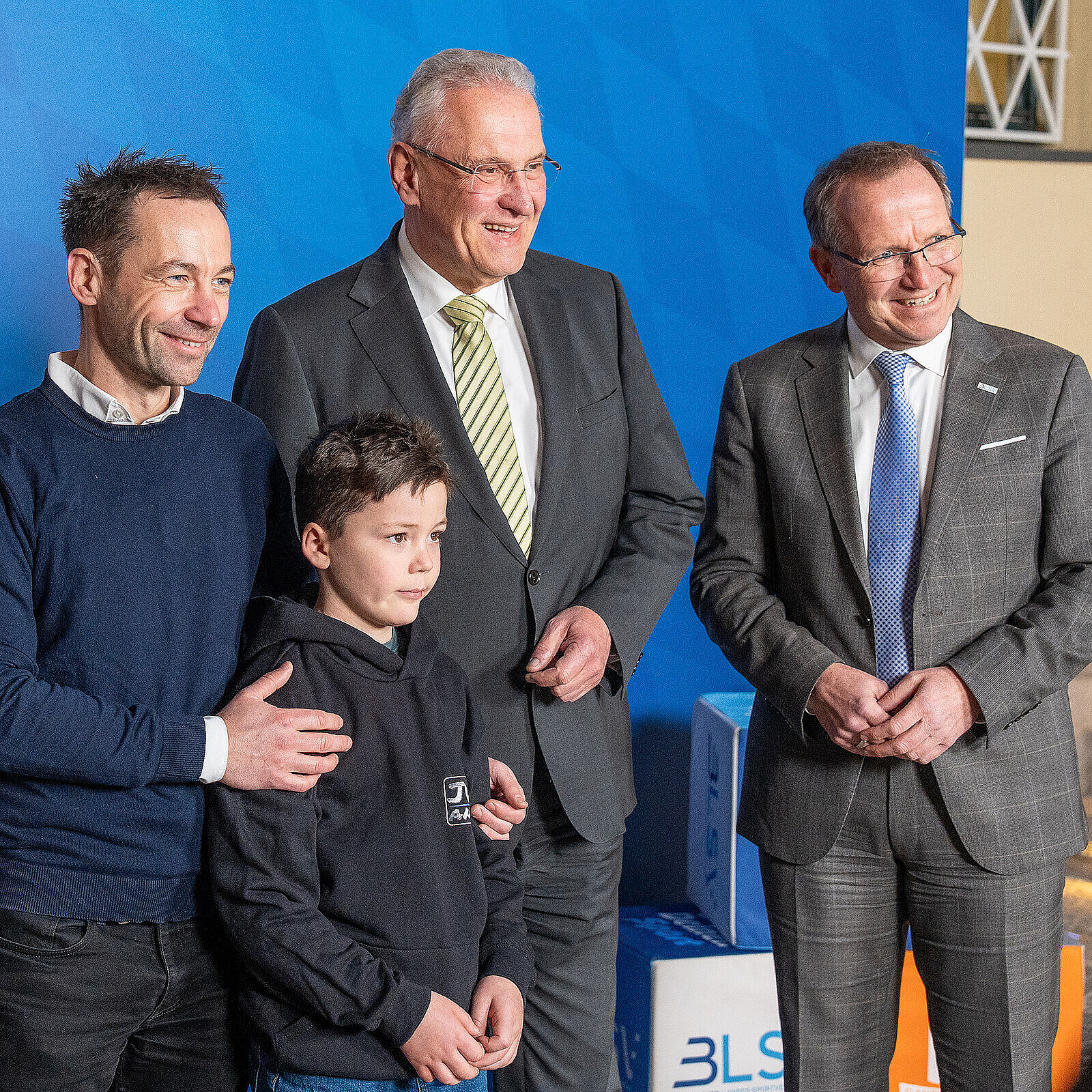 Sechs Personen (von re. nach li. Jörg Ammon, BLSV, Innenminister Joachim Herrmann, Emil Rucktäschel mit Vater) stehen in einer Reihe vor einer blauen Wand mit dem Schriftzug „Bayerisches Staatsministerium des Innern, für Sport und Integration“ und dem bayerischen Wappen. Zwei der Personen sind Kinder, die übrigen vier Erwachsene, von denen drei Anzüge tragen. Im Hintergrund sind Treppenstufen und einige Würfel mit Logos zu sehen.