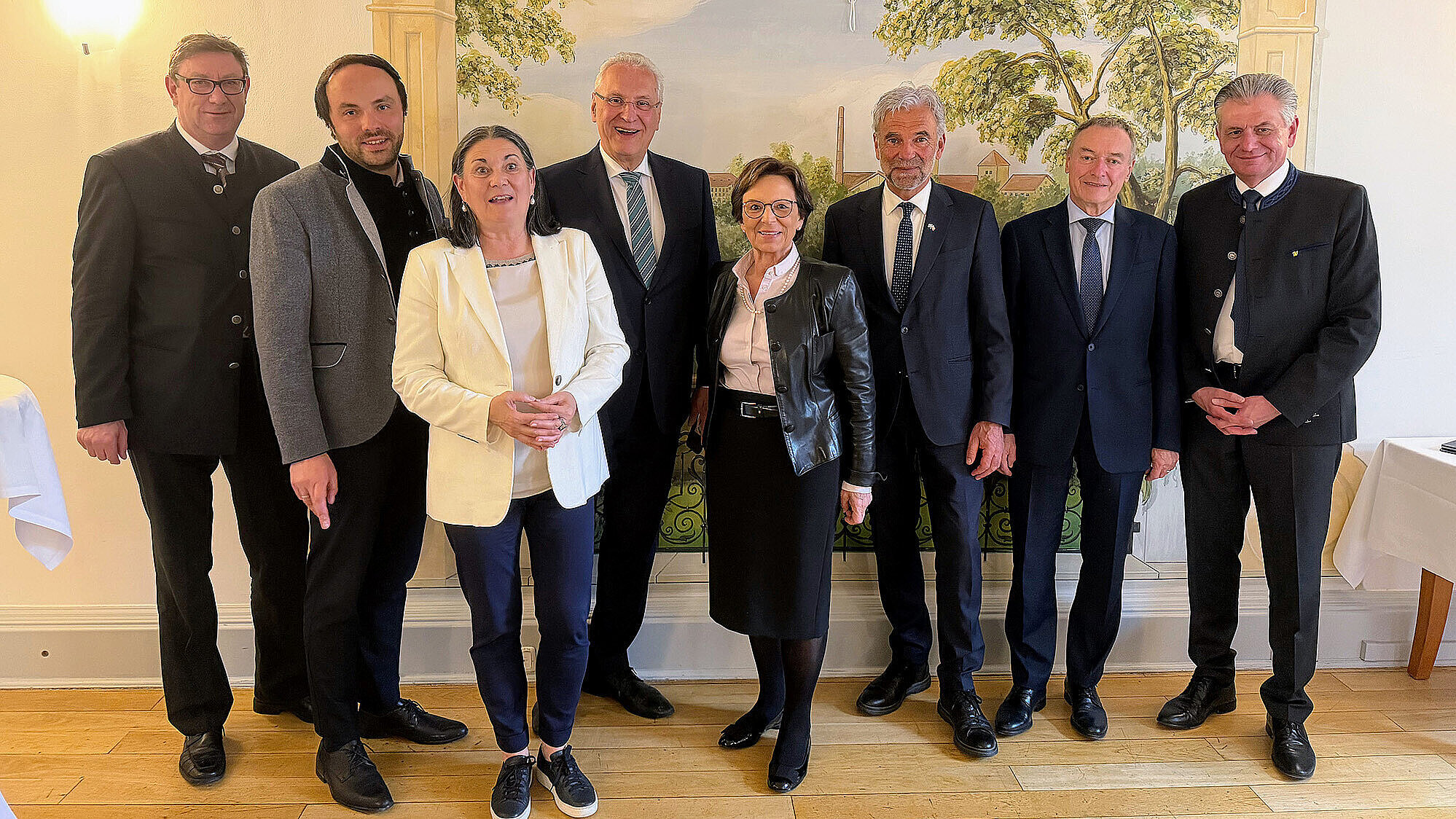 Acht Erwachsene stehen in einer Reihe vor einer dekorativen Wandmalerei in einem hellen Raum, alle formell oder halb-formell gekleidet.