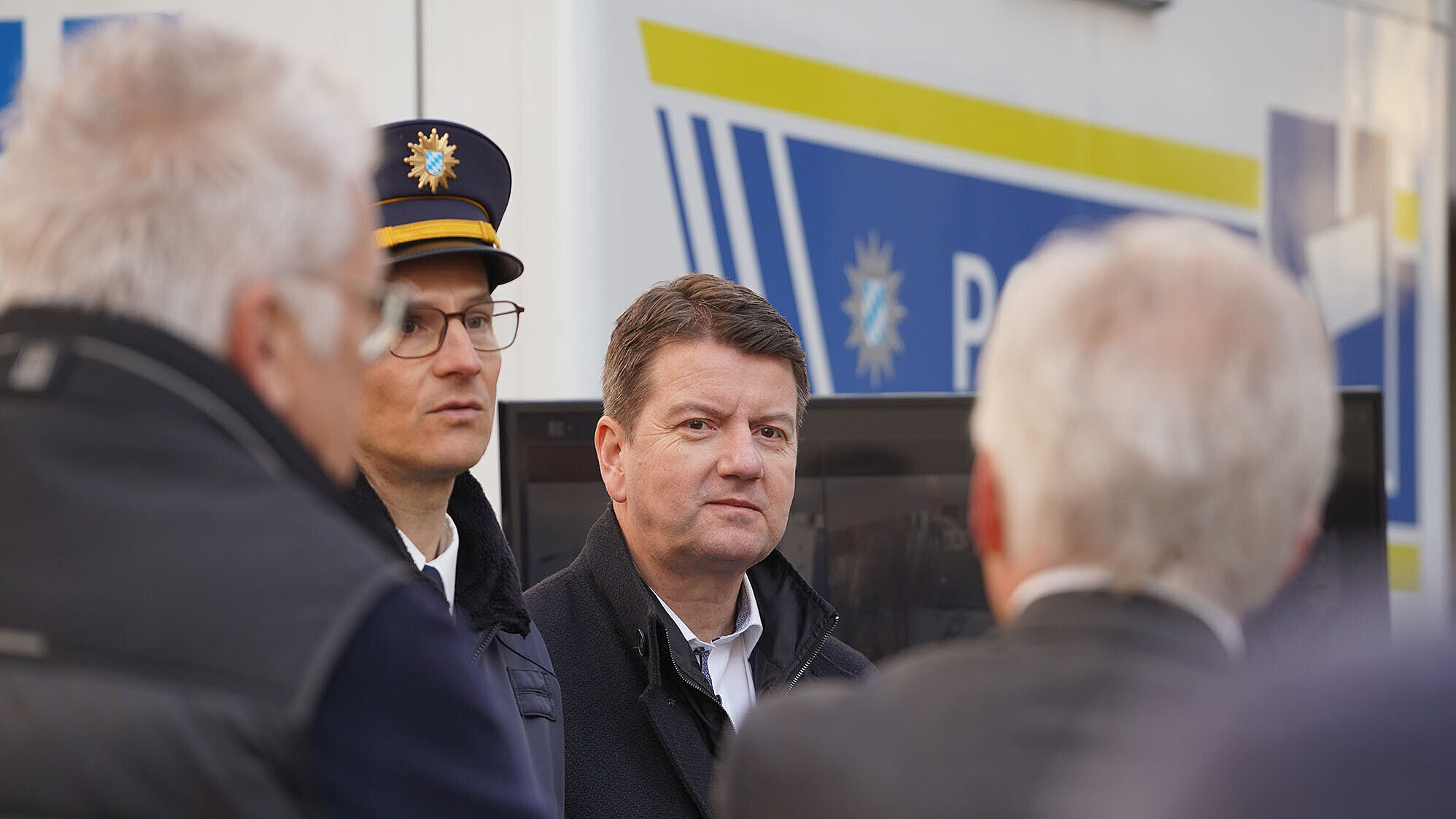 Vier Männer im Gespräch vor einem Polizeifahrzeug; einer trägt Polizeiuniform mit bayerischem Wappen, im Hintergrund die Aufschrift „Polizei“.