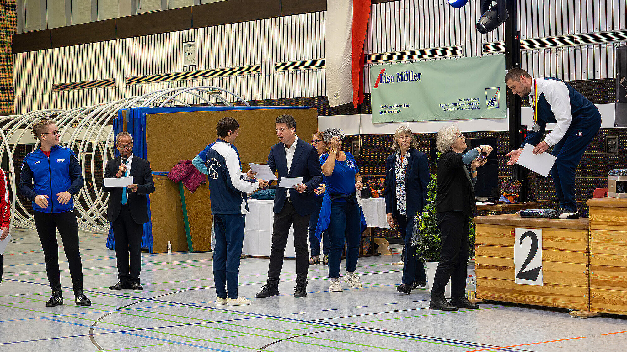 Das Bild zeigt eine Siegerehrung bei einer Sportveranstaltung. Mehrere Personen stehen in einer Halle, einige halten Urkunden, während eine Person auf der Bühne einem anderen die Hand schüttelt. Es ist eine feierliche Atmosphäre mit mehreren Teilnehmern und Zuschauern.