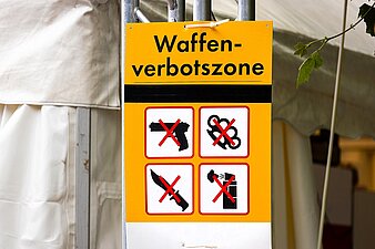 Schild Waffenverbotszone mit durchgestrichenen Symbolen von verschiedenen Waffen an einem Geländer befestigt