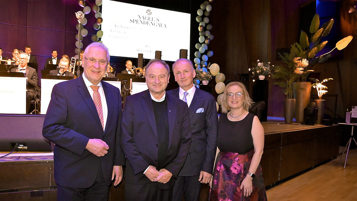 Vier Personen stehen in festlicher Kleidung nebeneinander (1.v.li. Innenminister Joachim Herrmann) in einem Saal vor einer Bühne, auf der eine Band spielt. Im Hintergrund ist eine große Leinwand mit der Aufschrift „Nägel’s Spendengala“ und eine Dekoration aus weißen und goldenen Luftballons zu sehen. Die Frau trägt ein schwarzes Oberteil und einen bunten Rock, die Männer Anzüge. Der Boden ist aus Holz, und im Raum sind Pflanzen und warme Beleuchtung vorhanden.