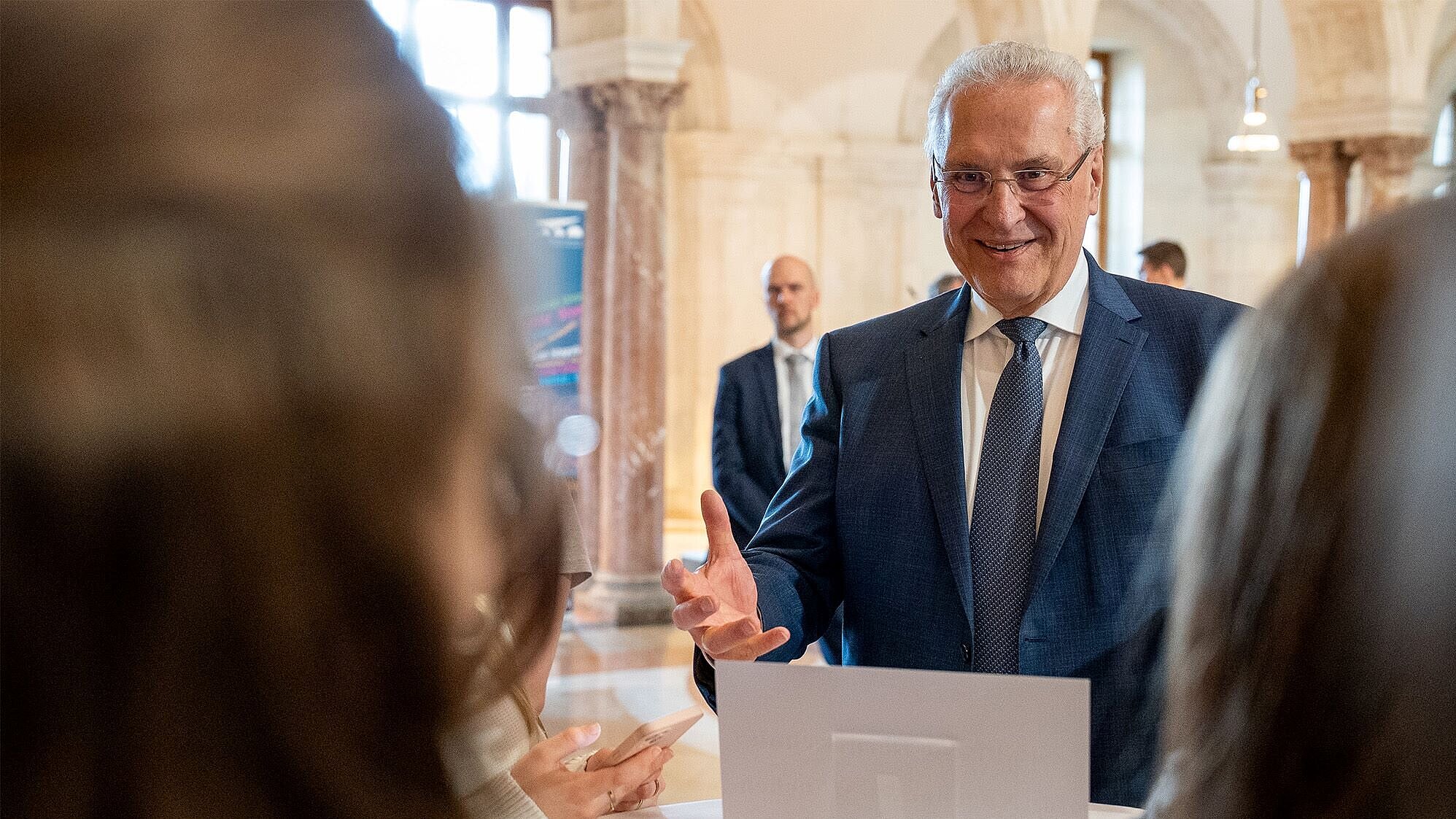 Auf dem Bild ist Innenminister Joachim Herrmann zu sehen, wie er sich freudig mit Schülerinnen und Schülern bei der Veranstaltung zum Lernort Staatsregierung unterhält.