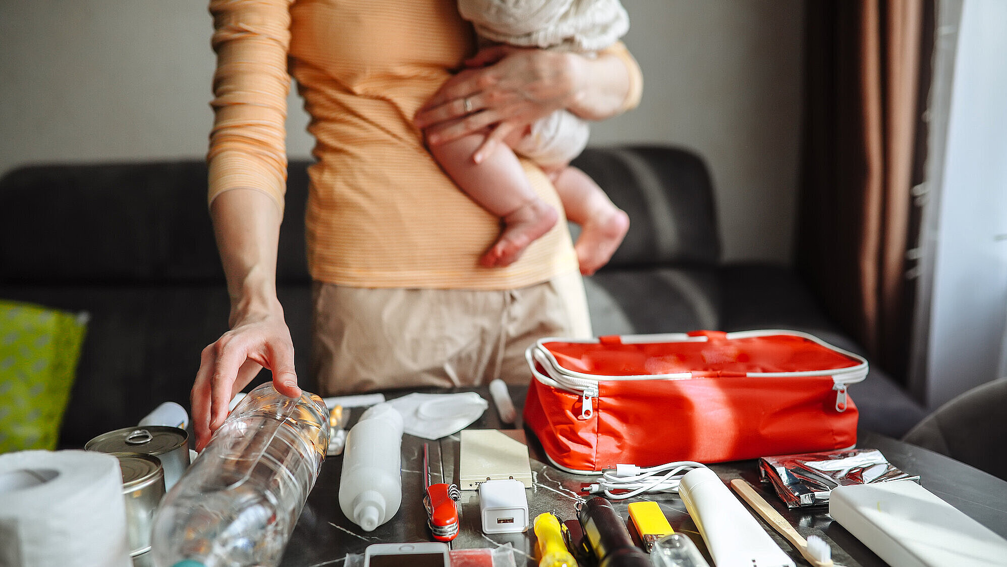 Mutter mit Baby-Mädchen packt Notfallset mit Medikamenten, Dokumenten, Wasser, Toilettenpapier, Taschenlampe, Messer, Telefon, Powerbank, Konserven und Wasser.