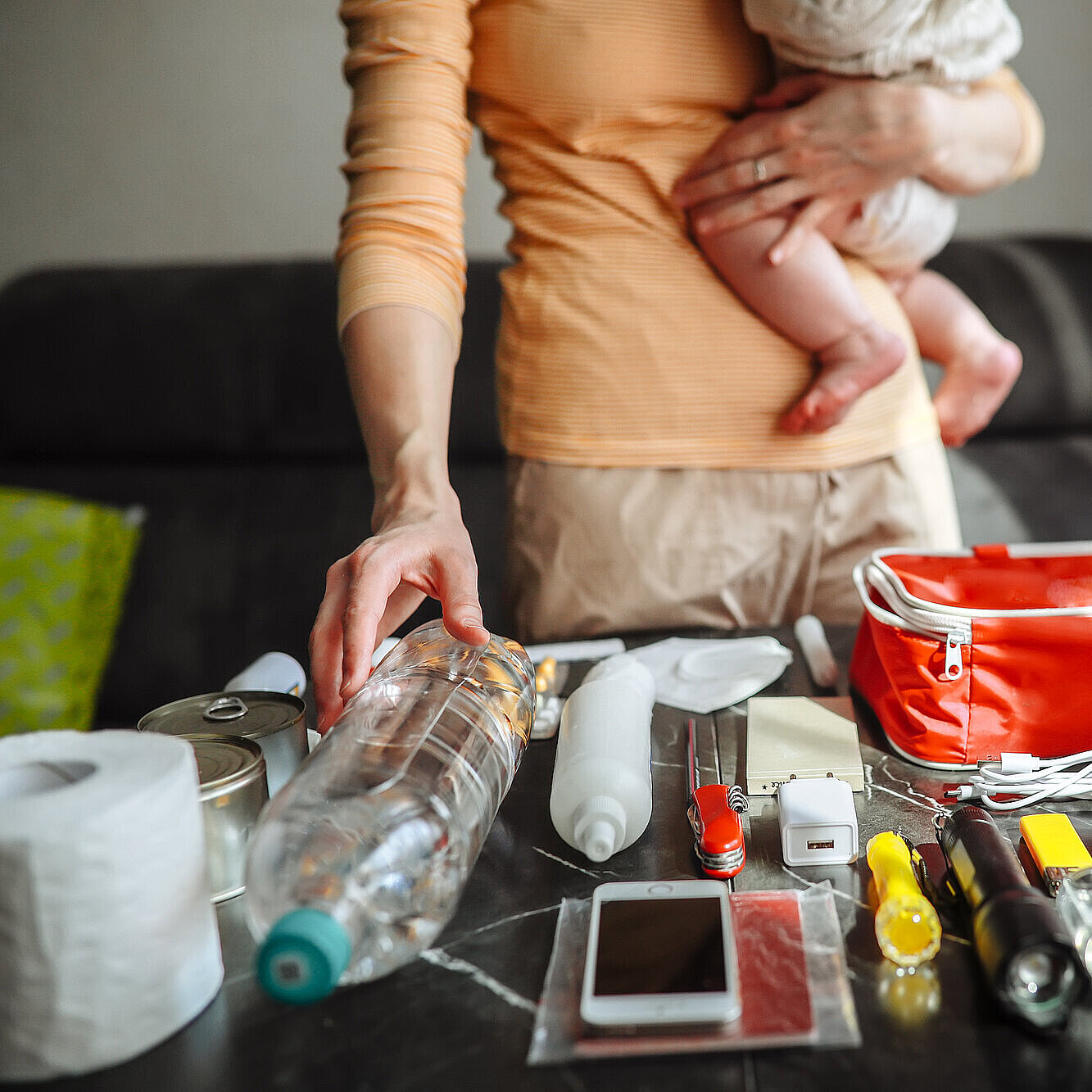 Mutter mit Baby-Mädchen packt Notfallset mit Medikamenten, Dokumenten, Wasser, Toilettenpapier, Taschenlampe, Messer, Telefon, Powerbank, Konserven und Wasser.