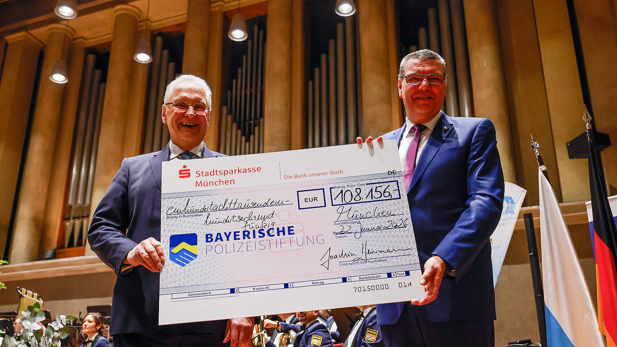 Zwei Männer in Anzügen stehen auf einer Bühne in einem Konzertsaal und halten gemeinsam einen großen symbolischen Spendenscheck der Stadtsparkasse München über 108.156 Euro zugunsten der Bayerischen Polizeistiftung. Im Hintergrund sind Musiker in Uniformen mit Blasinstrumenten zu sehen, sowie eine große Orgel und eine Bayerische Flagge.