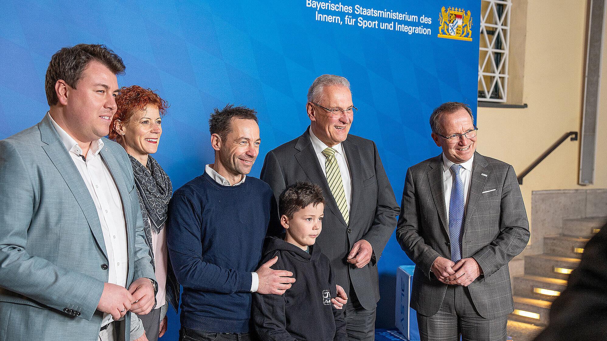 Sechs Personen (von re. nach li. Jörg Ammon, BLSV, Innenminister Joachim Herrmann, Emil Rucktäschel mit Vater) stehen in einer Reihe vor einer blauen Wand mit dem Schriftzug „Bayerisches Staatsministerium des Innern, für Sport und Integration“ und dem bayerischen Wappen. Zwei der Personen sind Kinder, die übrigen vier Erwachsene, von denen drei Anzüge tragen. Im Hintergrund sind Treppenstufen und einige Würfel mit Logos zu sehen.