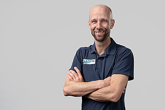 Pius Paschke vor hellem Hintergrund, trägt dunkles Poloshirt mit rechteckigem Aufnäher und hat die Arme verschränkt.