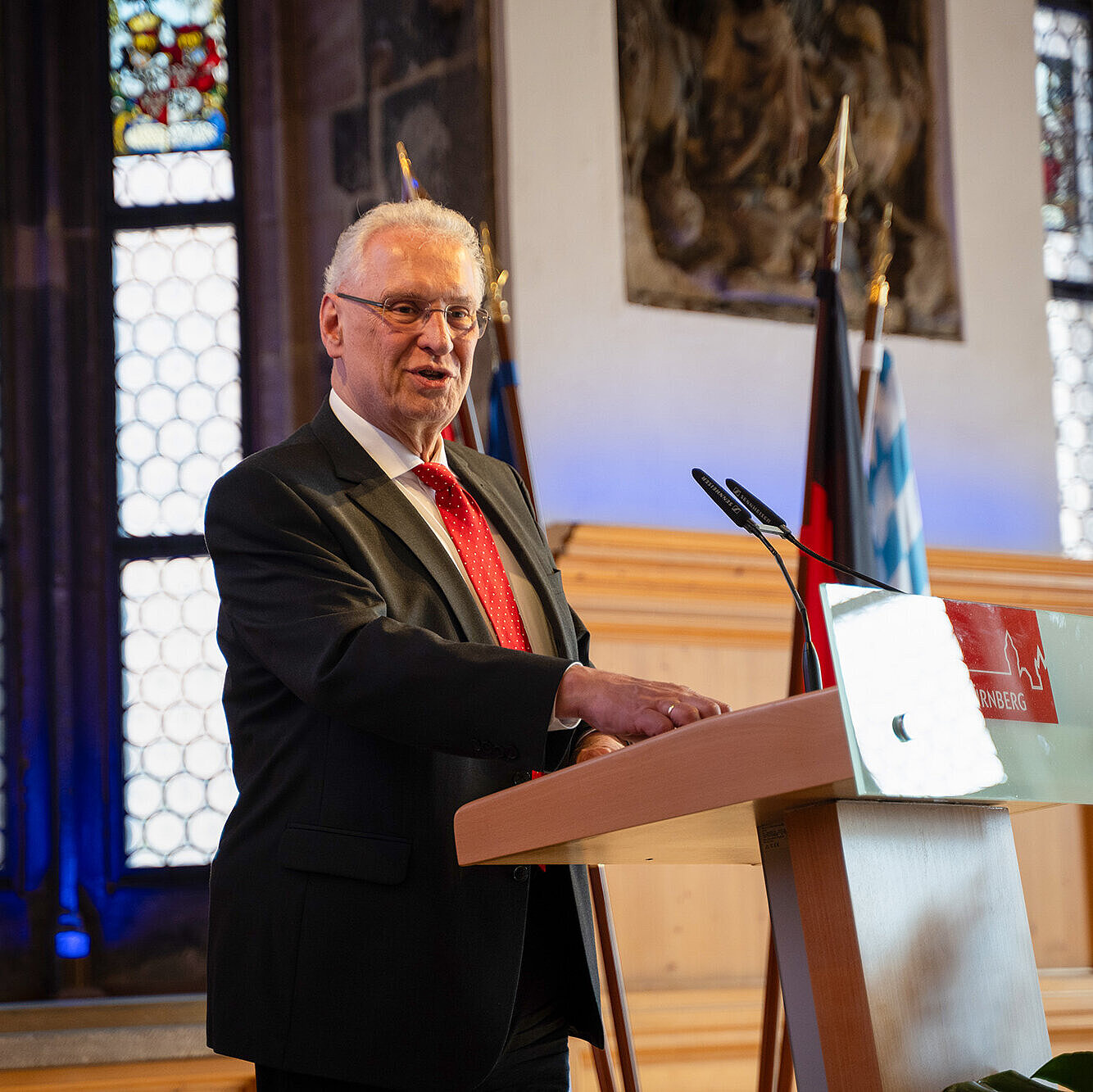 Mann in dunklem Anzug und roter Krawatte steht am Rednerpult, Gesicht unkenntlich gemacht; im Hintergrund hohe Fenster, Fahnen und Dekoration.
