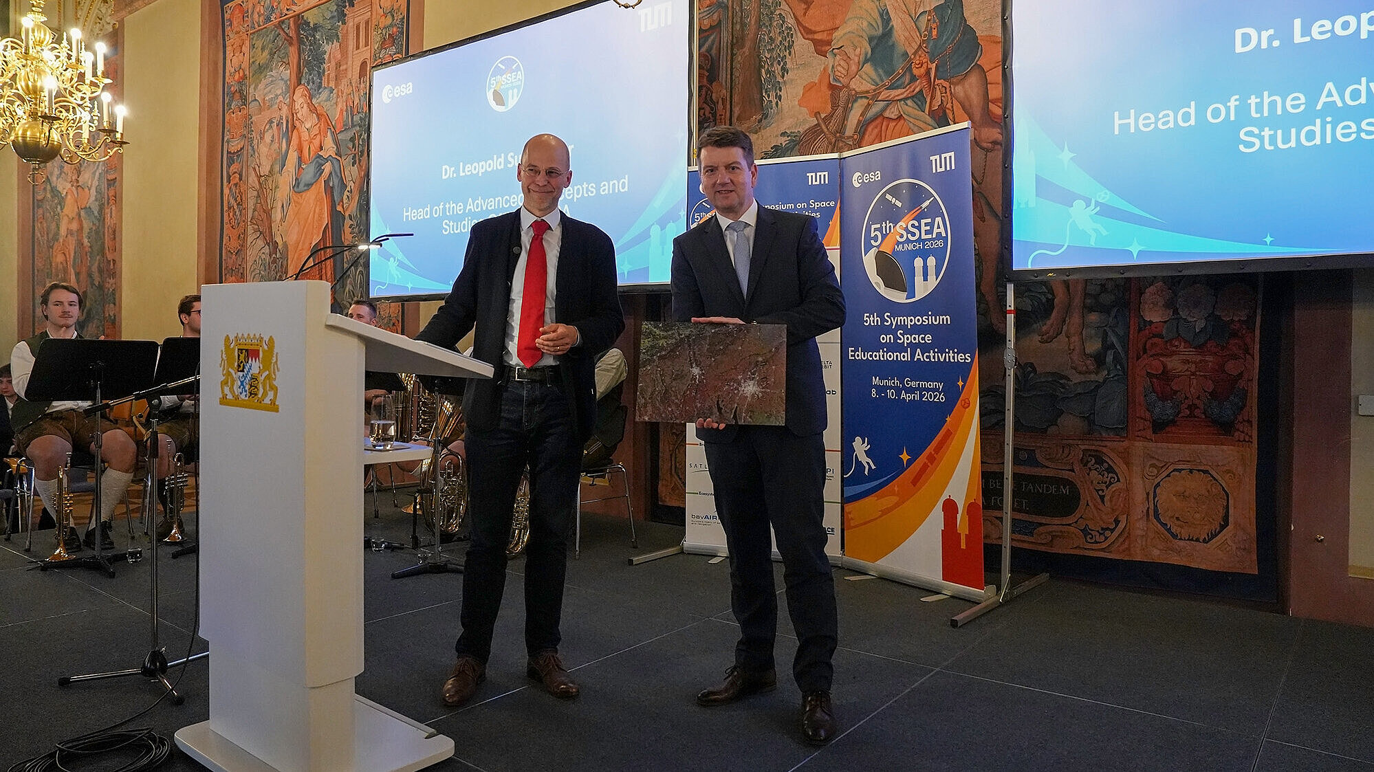 Zwei Personen stehen auf einer Bühne in einem prunkvollen Saal; eine Person spricht am Rednerpult, die andere hält eine Tafel, im Hintergrund Banner und Projektionsbildschirme.