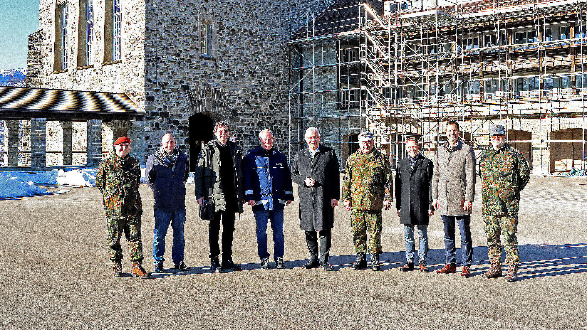 Eine Gruppe von neun Personen steht vor einem historischen Gebäude aus grobem Stein mit mehreren Fenstern und einem eingerüsteten Anbau. Drei der Personen tragen Tarnuniformen der Bundeswehr, die anderen sind in zivilen Mänteln und Jacken gekleidet. Im Hintergrund ist Schnee auf dem Boden zu sehen, und die Szene findet an einem sonnigen Tag statt.