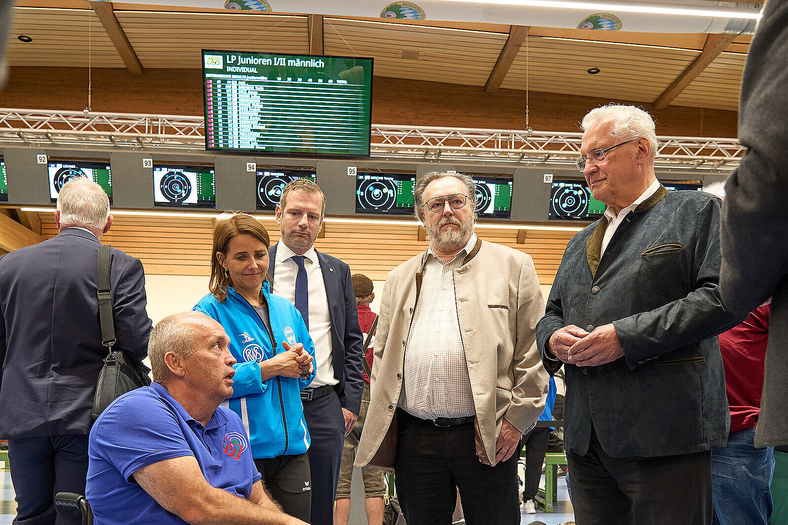 Ein Mann im Rollstuhl spricht mit drei stehenden Personen und Innenminister Joachim Herrmann (rechts im Bild)  in einem Wartebereich. Im Hintergrund sind Anzeigetafeln und weitere Personen zu sehen.