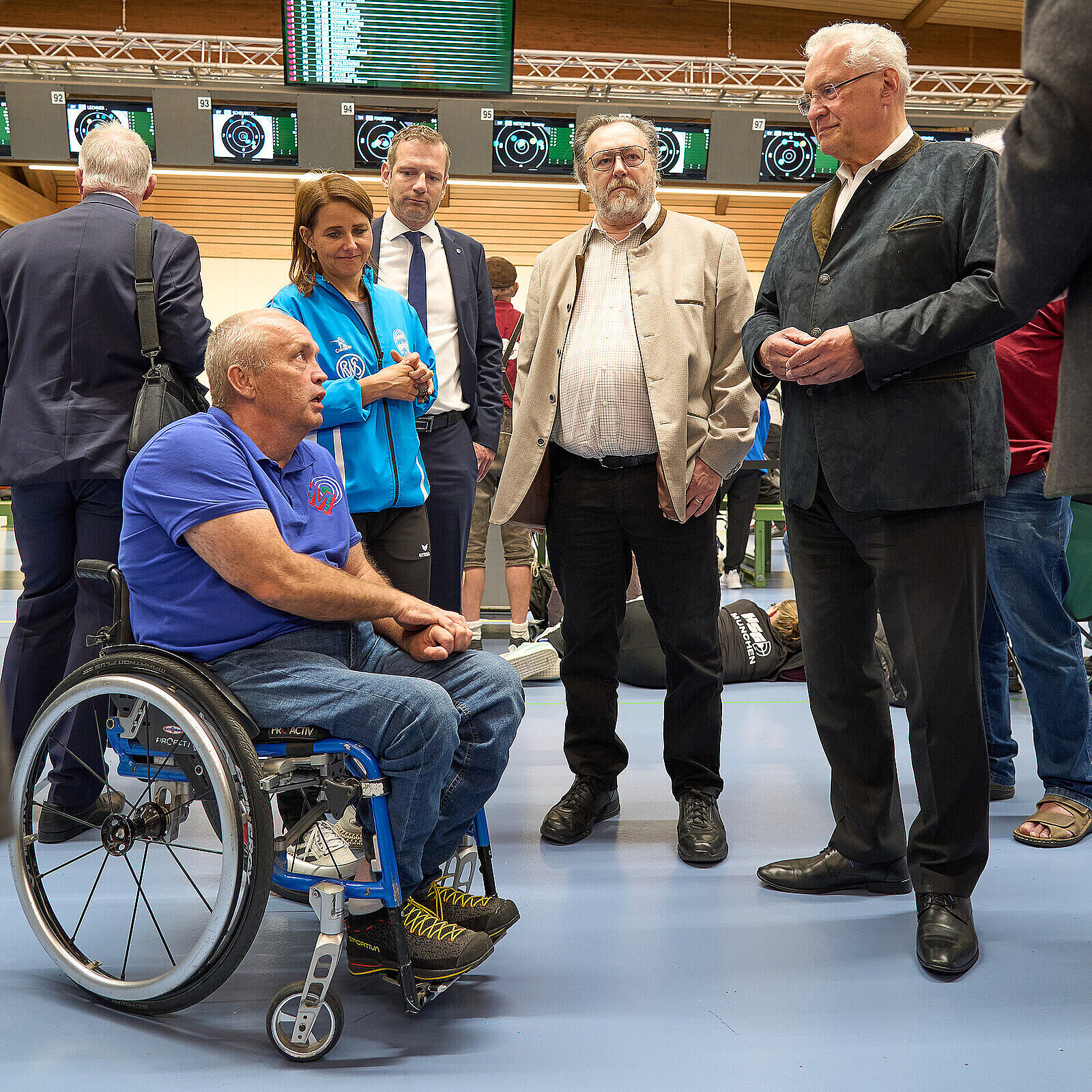 Ein Mann im Rollstuhl spricht mit drei stehenden Personen und Innenminister Joachim Herrmann (rechts im Bild)  in einem Wartebereich. Im Hintergrund sind Anzeigetafeln und weitere Personen zu sehen.