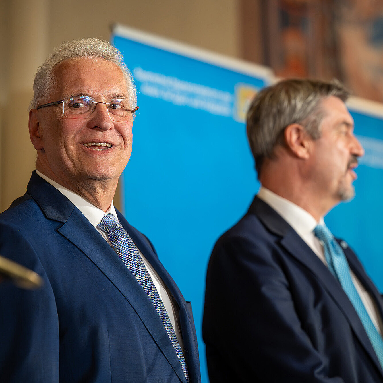 links Innenminister Joachim Herrmann, rechts Ministerpräsident Dr. Markus Söder mit Blick ins Publikum in der Reszidenz München, sie stehen vor einer blauen Pressewand