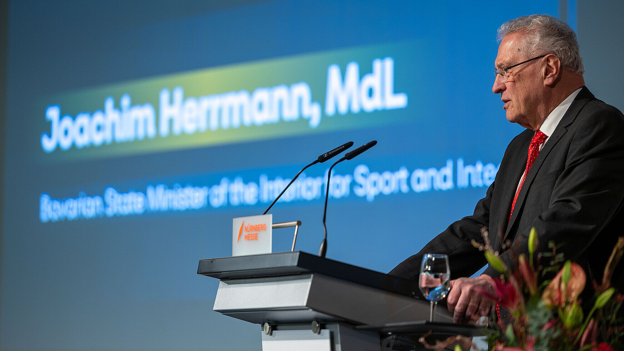 Innenminister Joachim Herrmann stehen am Rednerpult, links von ihm eine Bildschirmpräsentation mit seinem Namen.
