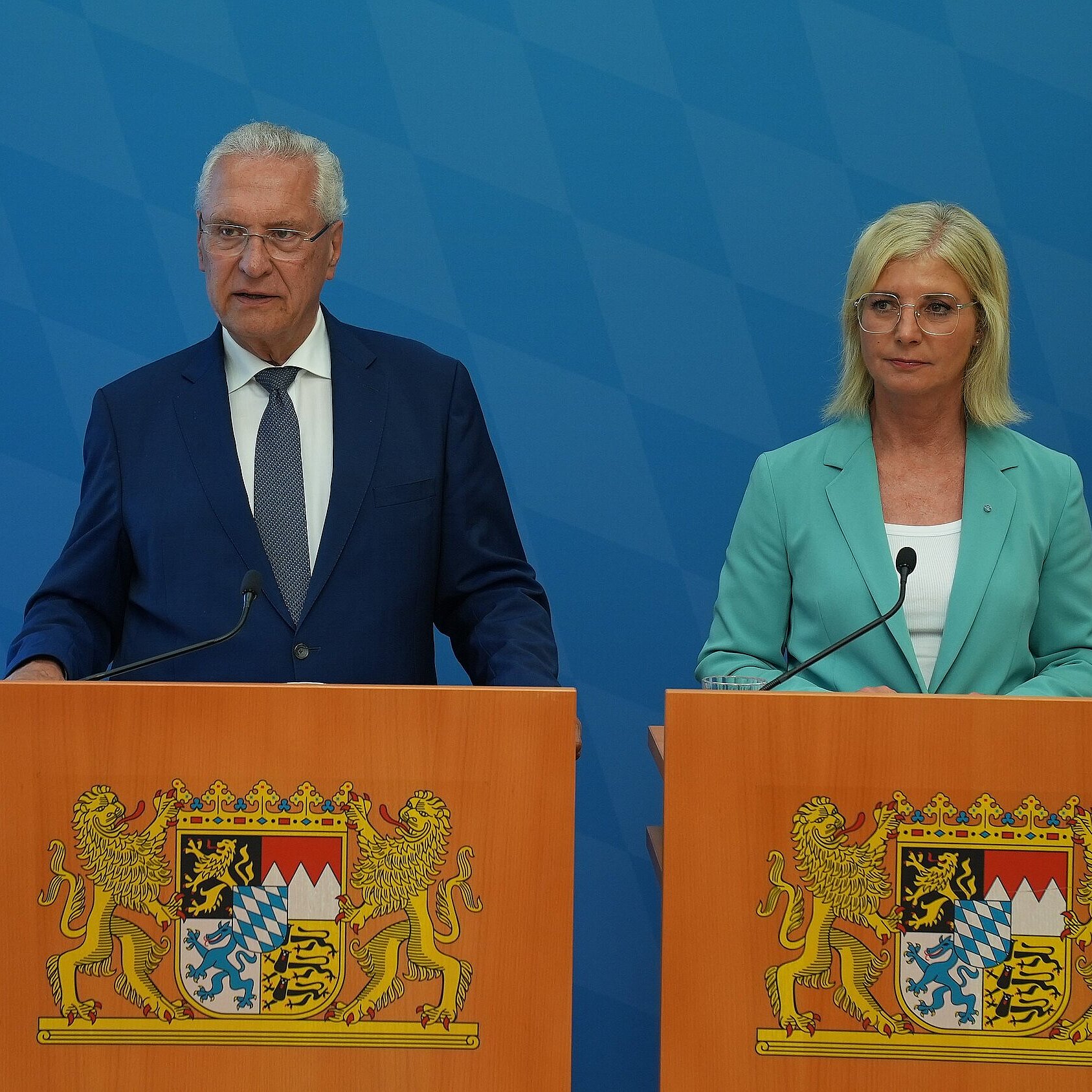Zwei Personen, eine Dame und ein Mann jeweils an einem Rednerpult, das Rednerpult ist in Holzoptik mit bayerischem Staatswappen