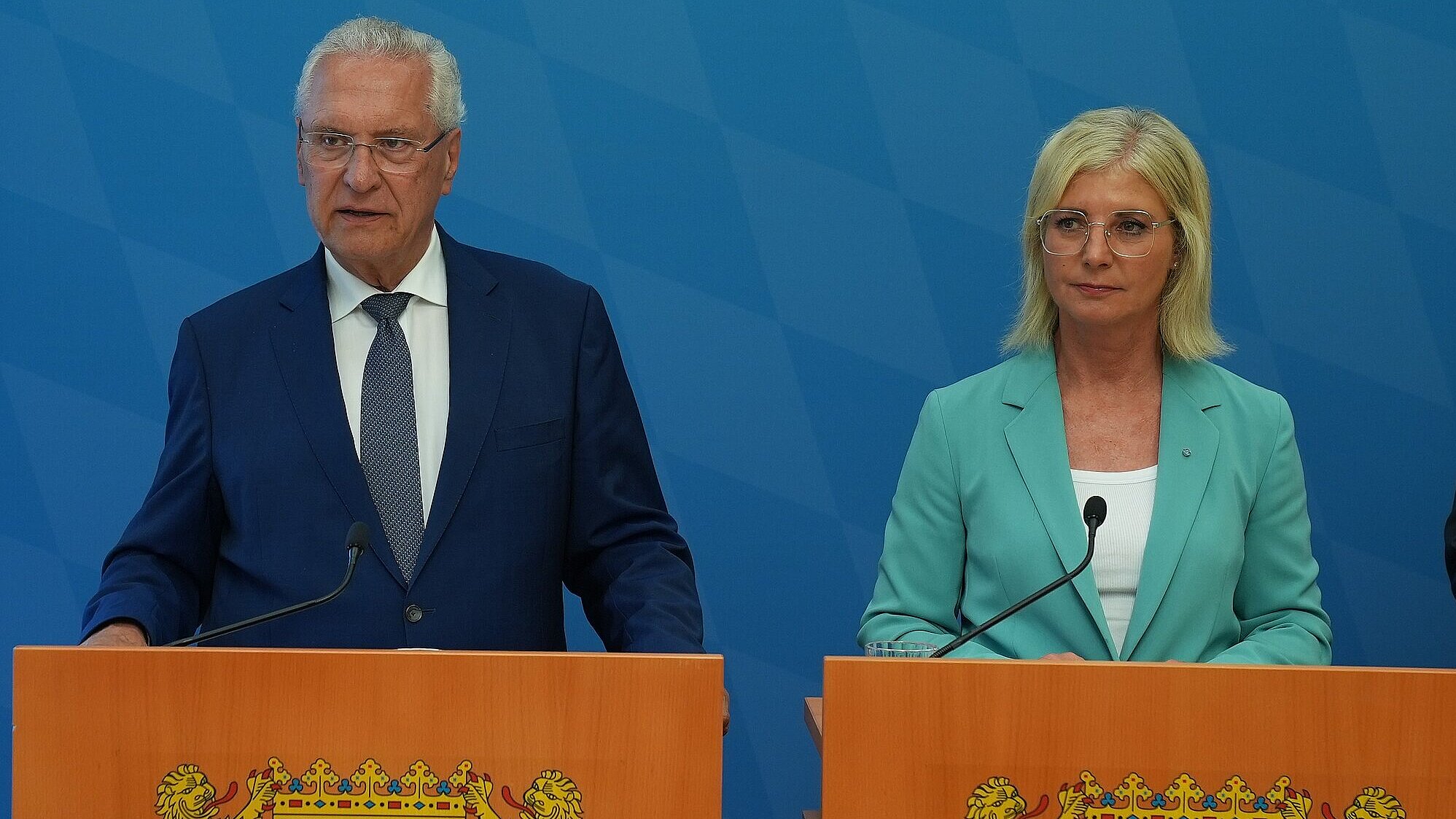 Zwei Personen, eine Dame und ein Mann jeweils an einem Rednerpult, das Rednerpult ist in Holzoptik mit bayerischem Staatswappen