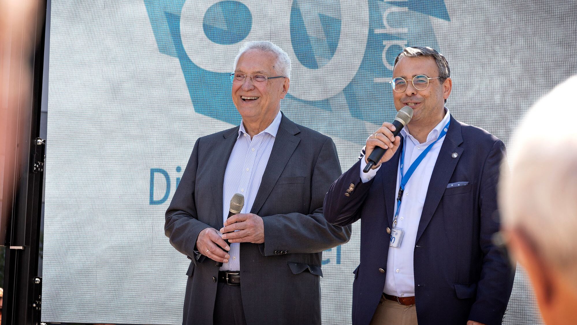 Innenminister Joachim Herrmann und BLKA-Präsident Norbert Radmacher lachend mit Mikrofonen auf einer Bühne. Hinter ihnen Bildschirmpräsentation mit Logo "80 Jahre Bayerische Polizei"