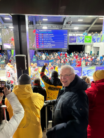 Innenminister Herrmann lächelt Kamera zu, hinter ihm jubelnde Menge und Ergebnisliste auf Bildschirm