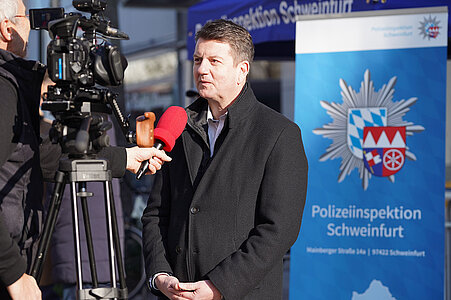 Männer im Gespräch vor einem Polizeifahrzeug; einer trägt Polizeiuniform mit bayerischem Wappen, im Hintergrund die Aufschrift „Polizei“.