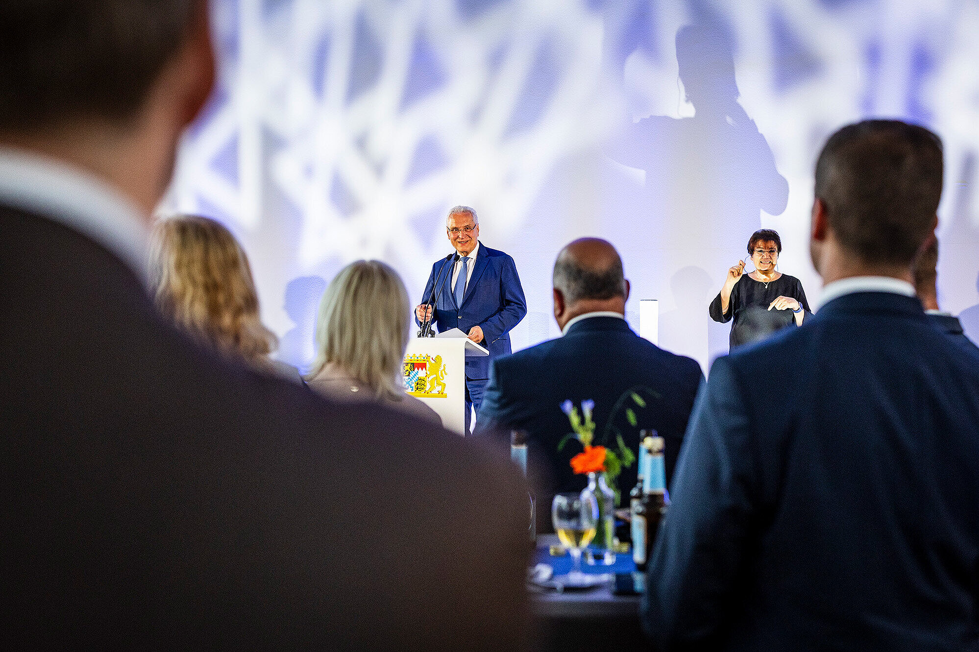 Innenminister Joachim Herrmann spricht bei der Eröffnung der Special Olympics Landesspiele in Erlangen am Rednerpult, im Hintergrund steht eine Gebärdensprachdolmetscherin. Im Vordergrund sind unscharf mehrere Zuhörerinnen und Zuhörer zu sehen, die ihm aufmerksam lauschen. Die festliche Atmosphäre wird durch Blumen, Getränke und Lichtreflexionen unterstrichen.