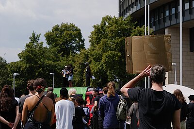 Menschenmenge, alle Personen sind von hinten abgebildet mit Blick auf eine Bühne, sie halten Schilder hoch 