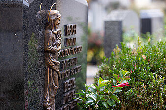 Grabstein mit einer bronzefarbenen Marienfigur umgeben von Pflanzen