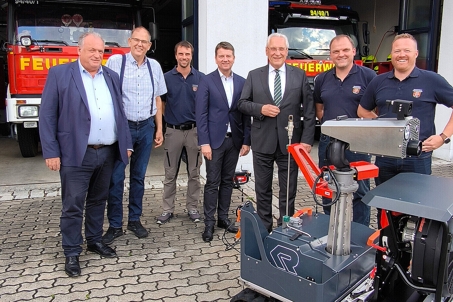 Gruppenfoto vor der Feuerwache Möhrendorf mit mehreren Einsatzfahrzeugen im Hintergrund. Sieben Männer, darunter Vertreter der Feuerwehr, Kommunalpolitik sowie Staatsminister Joachim Herrmann (3. von links) und Innenstaatssekretär Sandro Kirchner (4. von links), stehen nebeneinander und präsentieren einen modernen, ferngesteuerten Löschroboter auf Kettenfahrwerk. Die Szene spielt sich auf dem Hof vor der offenen Fahrzeughalle ab.