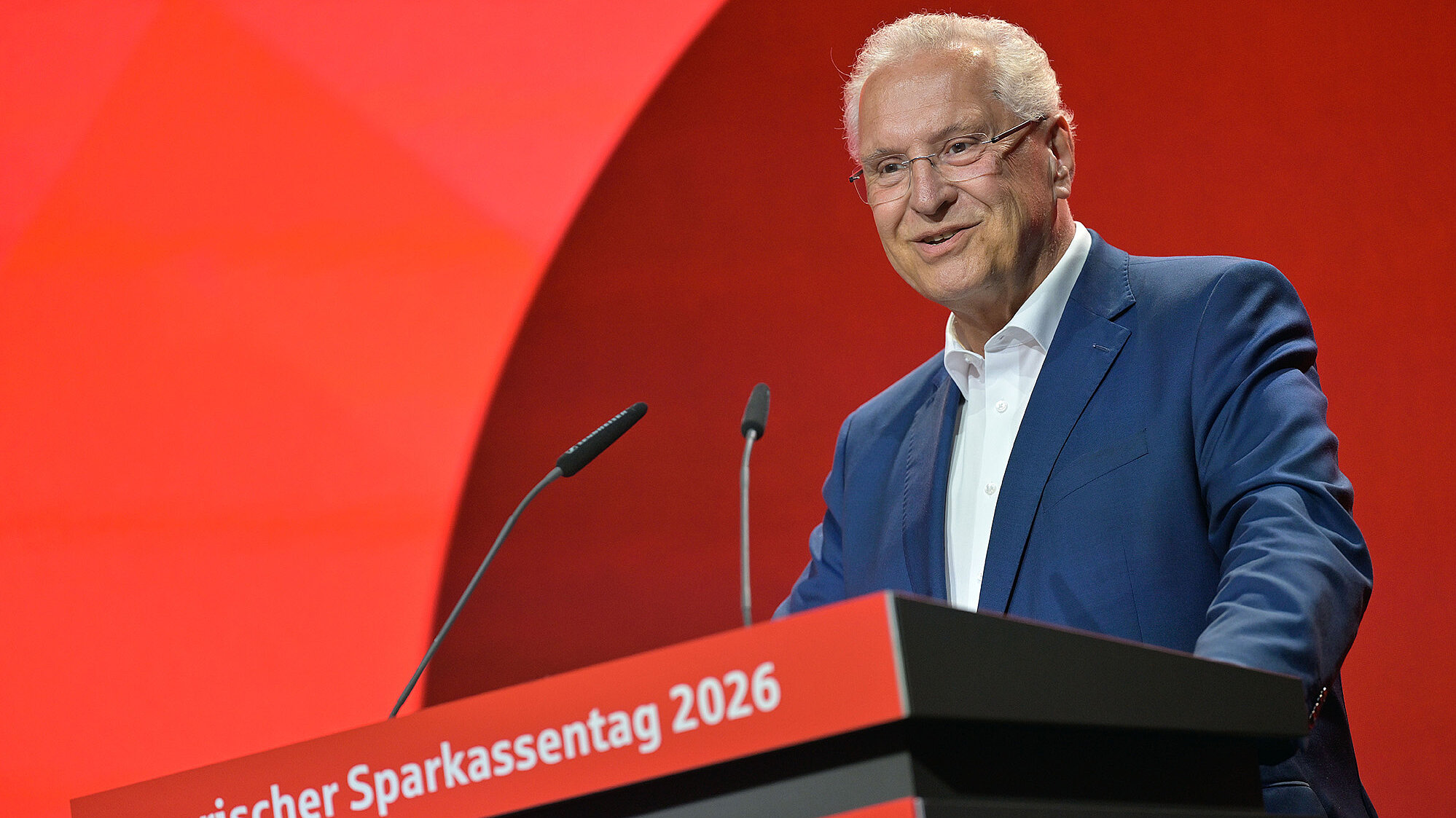Älterer Mann in dunklem Anzug und weißem Hemd steht lächelnd an einem roten Rednerpult mit der Aufschrift „Bayerischer Sparkassentag 2026“. Zwei Mikrofone sind sichtbar, im Hintergrund eine rote Bühnenkulisse.