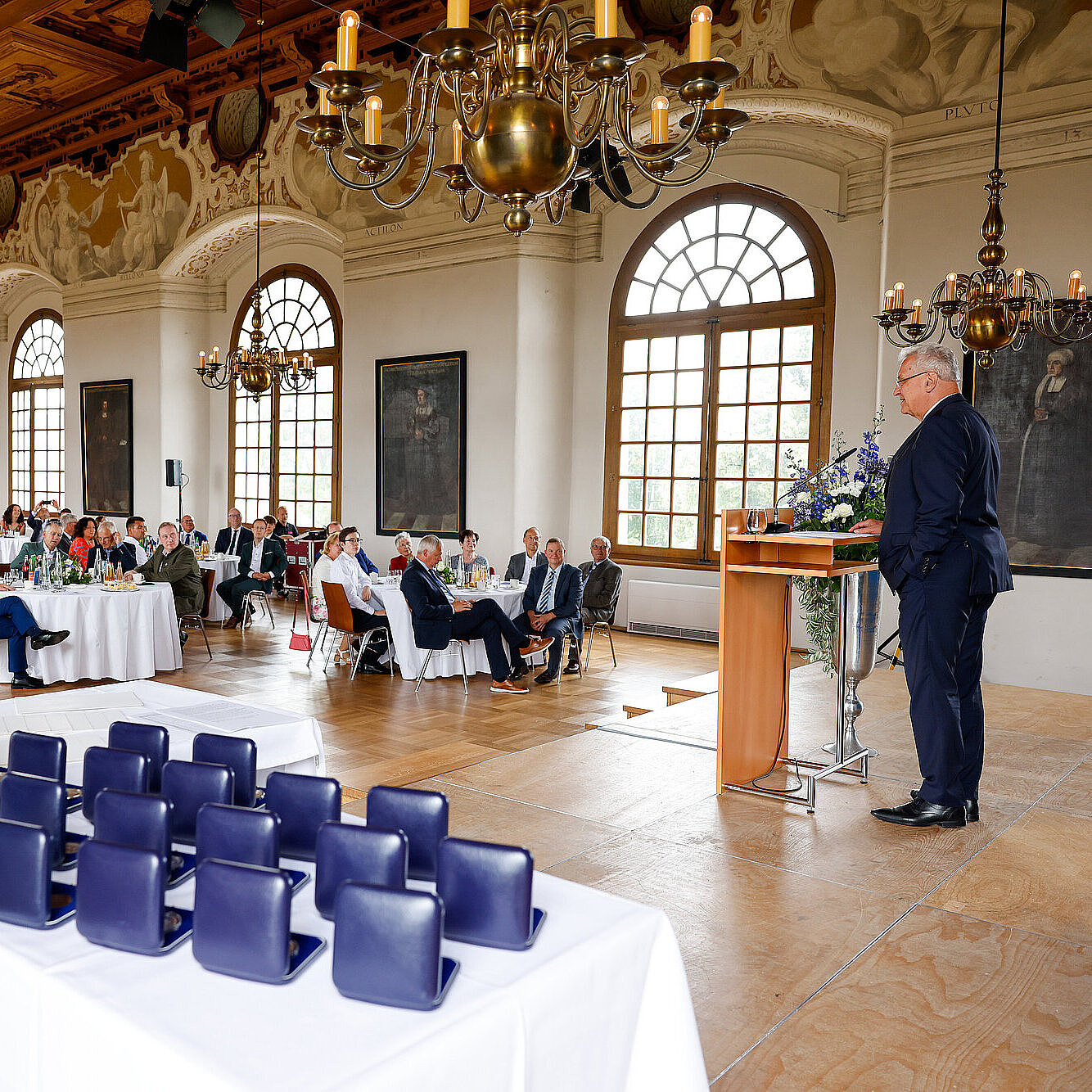 Blick in den Festsaal, rechts Innenminister Herrmann am Rednerpult, links Publikum in Stuhlreihen, im Vordergrund kommunale Verdienstmedaillen auf Tisch in Schachteln